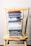 A stack of colorful books is neatly arranged on a worn yellow wooden chair, set against a white brick wall. The books vary in size and color, with titles related to faith, wisdom, and encouragement.