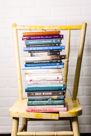 A stack of colorful books is neatly arranged on a worn yellow wooden chair, set against a white brick wall. The books vary in size and color, with titles related to faith, wisdom, and encouragement.