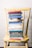 A stack of colorful books is neatly arranged on a worn yellow wooden chair, set against a white brick wall. The books vary in size and color, with titles related to faith, wisdom, and encouragement.