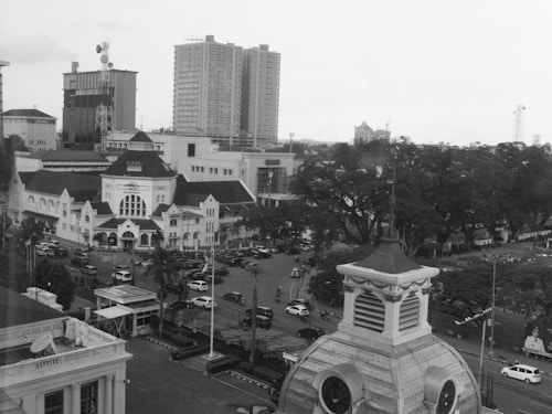 An urban scene featuring historic and modern architecture. A prominent older building with classic design elements is in the foreground, surrounded by busy streets filled with cars and occasional pedestrians. In the background, modern high-rise buildings and some trees can be seen.