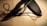 A knife with a serrated edge is placed on a wooden surface. It is accompanied by a black sheath and a black cord looped through a metal eyelet on the sheath.