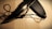 A knife with a serrated edge is placed on a wooden surface. It is accompanied by a black sheath and a black cord looped through a metal eyelet on the sheath.