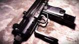 A close-up view of a firearm, resting on a textured surface. The weapon has a sleek design with multiple grooves on the handle, and the lighting casts dramatic shadows.