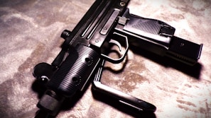 A close-up view of a firearm, resting on a textured surface. The weapon has a sleek design with multiple grooves on the handle, and the lighting casts dramatic shadows.
