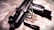 A close-up view of a firearm, resting on a textured surface. The weapon has a sleek design with multiple grooves on the handle, and the lighting casts dramatic shadows.