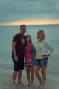 A family of three stands on a beach with the ocean in the background. The sky is overcast with a hint of sunset on the horizon. The adults are smiling, and the young girl stands between them. The sand and water create a serene backdrop.