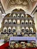 An ornate altar is adorned with a series of religious statues, each placed within decorative niches. The altar features gold and intricate carvings with religious iconography. At the forefront, a white altar cloth embroidered with floral patterns covers a table surrounded by colorful flowers. Above, there are elegant chandeliers and artistic frescoes on the arched ceiling, depicting various religious scenes.