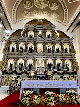 An ornate altar is adorned with a series of religious statues, each placed within decorative niches. The altar features gold and intricate carvings with religious iconography. At the forefront, a white altar cloth embroidered with floral patterns covers a table surrounded by colorful flowers. Above, there are elegant chandeliers and artistic frescoes on the arched ceiling, depicting various religious scenes.