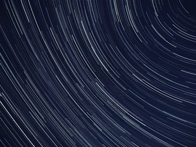A long-exposure photograph capturing the motion of stars in the night sky, creating circular streaks of light across a dark backdrop. The lines are mostly white with hints of blue, indicating the movement of stars across time.