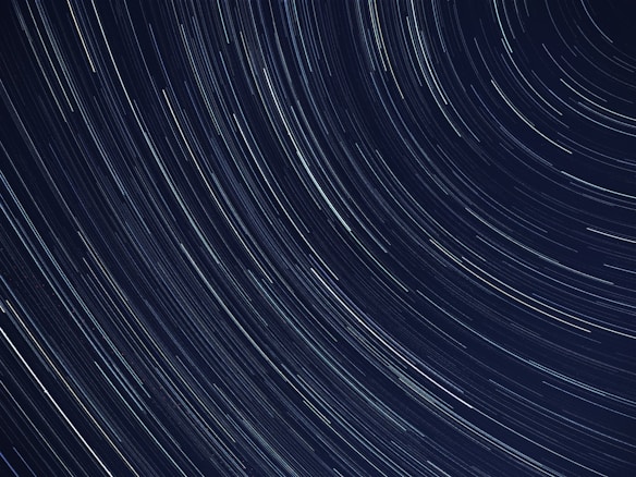 A long-exposure photograph capturing the motion of stars in the night sky, creating circular streaks of light across a dark backdrop. The lines are mostly white with hints of blue, indicating the movement of stars across time.