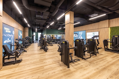 A modern gym interior with various exercise machines neatly arranged on a wooden floor. The walls are lined with motivational posters and parts are adorned with artificial greenery. Ceiling lights illuminate the space, highlighting the sleek and organized look of the equipment.
