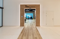 A modern fitness center entrance with wooden framing and the sign 'PRIME FITNESS' above the doorway. The interior features a spacious gym area with workout equipment visible and a motivational banner on the back wall. The flooring transitions from glossy tiles to a wood finish leading into the facility.
