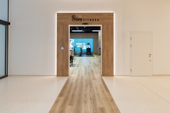 A modern fitness center entrance with wooden framing and the sign 'PRIME FITNESS' above the doorway. The interior features a spacious gym area with workout equipment visible and a motivational banner on the back wall. The flooring transitions from glossy tiles to a wood finish leading into the facility.