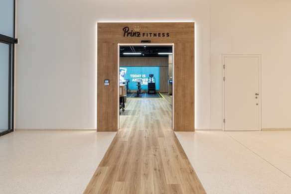 A modern fitness center entrance with wooden framing and the sign 'PRIME FITNESS' above the doorway. The interior features a spacious gym area with workout equipment visible and a motivational banner on the back wall. The flooring transitions from glossy tiles to a wood finish leading into the facility.
