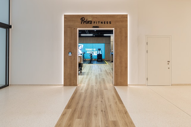 A modern fitness center entrance with wooden framing and the sign 'PRIME FITNESS' above the doorway. The interior features a spacious gym area with workout equipment visible and a motivational banner on the back wall. The flooring transitions from glossy tiles to a wood finish leading into the facility.