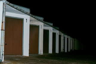 A row of closed garage doors with numbers from 1 to 8, illuminated by artificial light. The garages are aligned under a continuous sloped roof, and the setting appears to be nighttime, as the background is entirely dark.