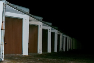 A row of closed garage doors with numbers from 1 to 8, illuminated by artificial light. The garages are aligned under a continuous sloped roof, and the setting appears to be nighttime, as the background is entirely dark.