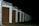 A row of closed garage doors with numbers from 1 to 8, illuminated by artificial light. The garages are aligned under a continuous sloped roof, and the setting appears to be nighttime, as the background is entirely dark.
