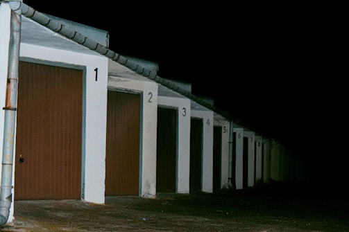 A row of closed garage doors with numbers from 1 to 8, illuminated by artificial light. The garages are aligned under a continuous sloped roof, and the setting appears to be nighttime, as the background is entirely dark.