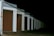 A row of closed garage doors with numbers from 1 to 8, illuminated by artificial light. The garages are aligned under a continuous sloped roof, and the setting appears to be nighttime, as the background is entirely dark.