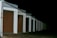 A row of closed garage doors with numbers from 1 to 8, illuminated by artificial light. The garages are aligned under a continuous sloped roof, and the setting appears to be nighttime, as the background is entirely dark.