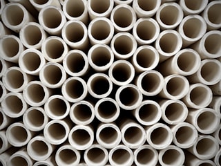 A close-up view of a large stack of uniform, cylindrical pipes arranged in a tightly packed, orderly pattern. The pipes are aligned vertically, creating a repetitive geometric design with circular openings visible.