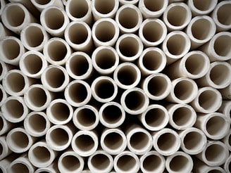 A close-up view of a large stack of uniform, cylindrical pipes arranged in a tightly packed, orderly pattern. The pipes are aligned vertically, creating a repetitive geometric design with circular openings visible.