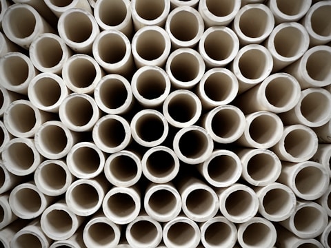 A close-up view of a large stack of uniform, cylindrical pipes arranged in a tightly packed, orderly pattern. The pipes are aligned vertically, creating a repetitive geometric design with circular openings visible.