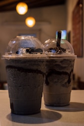 Two large plastic cups with dome lids filled with blended chocolate drinks and topped with whipped cream and chocolate sprinkles. The background is softly lit with warm overhead lights adding a cozy ambiance.