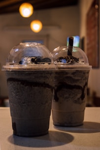 Two large plastic cups with dome lids filled with blended chocolate drinks and topped with whipped cream and chocolate sprinkles. The background is softly lit with warm overhead lights adding a cozy ambiance.