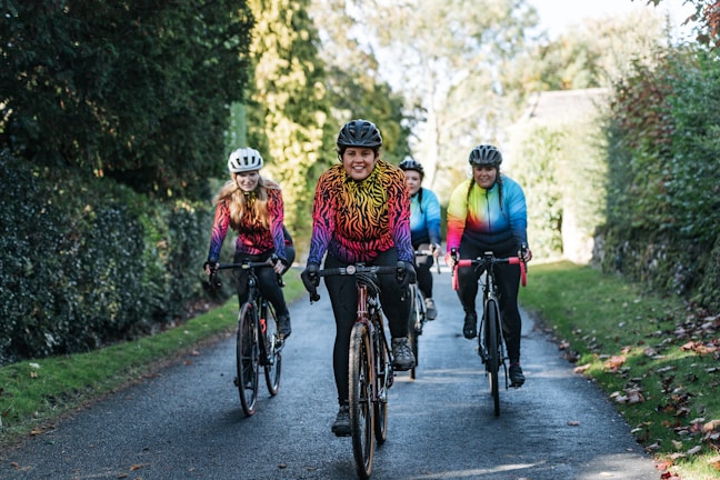 Four people riding bicycles on a paved road surrounded by lush greenery. They are all wearing vibrant, multicolored cycling jerseys and helmets. The group appears to be enjoying a scenic ride amidst nature on a sunny day.