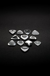 A set of eleven metallic and glossy guitar picks arranged neatly on a dark, textured surface. Each pick has a unique design, with varying patterns and finishes, suggesting precision craftsmanship and a focus on aesthetics.