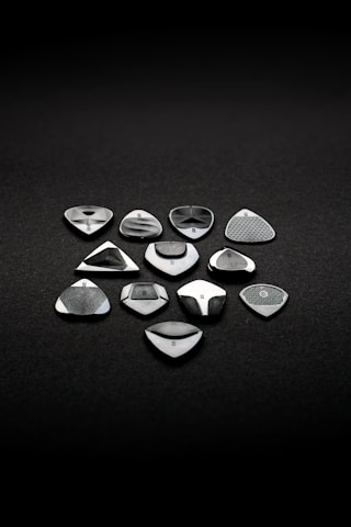 A set of eleven metallic and glossy guitar picks arranged neatly on a dark, textured surface. Each pick has a unique design, with varying patterns and finishes, suggesting precision craftsmanship and a focus on aesthetics.