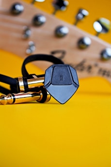 A close-up of a guitar headstock with tuning pegs, accompanied by a black guitar pick and two gold guitar jacks resting on a bright yellow background.