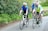 Three cyclists are riding on a road surrounded by green foliage. They are wearing helmets and colorful cycling gear, with two in blue jerseys and one in a yellow vest. The mood appears friendly and relaxed, as they are engaged in conversation while cycling.