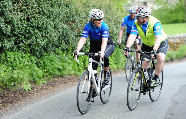 Three cyclists are riding on a road surrounded by green foliage. They are wearing helmets and colorful cycling gear, with two in blue jerseys and one in a yellow vest. The mood appears friendly and relaxed, as they are engaged in conversation while cycling.