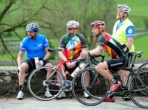 Four cyclists dressed in colorful cycling gear are gathered outdoors, each wearing helmets. Two are sitting on a stone wall while the other two are standing, holding their bikes. The background is a lush, green natural setting with trees and open space.