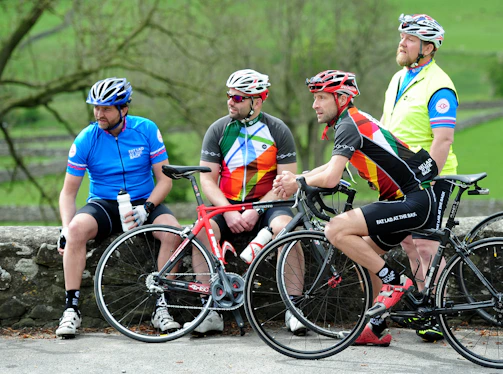 Four cyclists dressed in colorful cycling gear are gathered outdoors, each wearing helmets. Two are sitting on a stone wall while the other two are standing, holding their bikes. The background is a lush, green natural setting with trees and open space.
