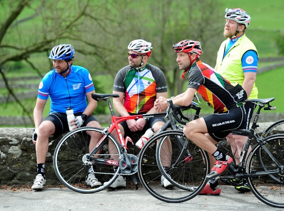 Four cyclists dressed in colorful cycling gear are gathered outdoors, each wearing helmets. Two are sitting on a stone wall while the other two are standing, holding their bikes. The background is a lush, green natural setting with trees and open space.