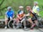 Four cyclists dressed in colorful cycling gear are gathered outdoors, each wearing helmets. Two are sitting on a stone wall while the other two are standing, holding their bikes. The background is a lush, green natural setting with trees and open space.