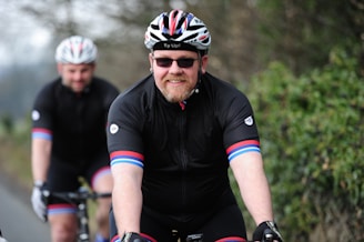 Two cyclists wearing matching black cycling gear and helmets ride along a road bordered by greenery. The cyclist in front has a beard and sunglasses and appears to be smiling.