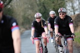 Several cyclists wearing matching black cycling outfits with red and blue stripes are riding on a road through a rural area. They are all wearing helmets and sunglasses. The path they are on is bordered by trees and bushes, creating a natural setting.