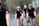 Several cyclists wearing matching black cycling outfits with red and blue stripes are riding on a road through a rural area. They are all wearing helmets and sunglasses. The path they are on is bordered by trees and bushes, creating a natural setting.