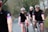 Several cyclists wearing matching black cycling outfits with red and blue stripes are riding on a road through a rural area. They are all wearing helmets and sunglasses. The path they are on is bordered by trees and bushes, creating a natural setting.