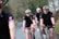 Several cyclists wearing matching black cycling outfits with red and blue stripes are riding on a road through a rural area. They are all wearing helmets and sunglasses. The path they are on is bordered by trees and bushes, creating a natural setting.