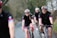 Several cyclists wearing matching black cycling outfits with red and blue stripes are riding on a road through a rural area. They are all wearing helmets and sunglasses. The path they are on is bordered by trees and bushes, creating a natural setting.
