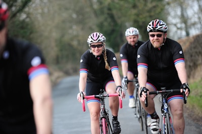 Several cyclists wearing matching black cycling outfits with red and blue stripes are riding on a road through a rural area. They are all wearing helmets and sunglasses. The path they are on is bordered by trees and bushes, creating a natural setting.