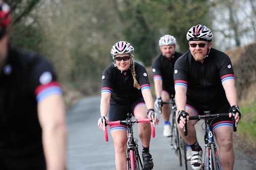 Several cyclists wearing matching black cycling outfits with red and blue stripes are riding on a road through a rural area. They are all wearing helmets and sunglasses. The path they are on is bordered by trees and bushes, creating a natural setting.