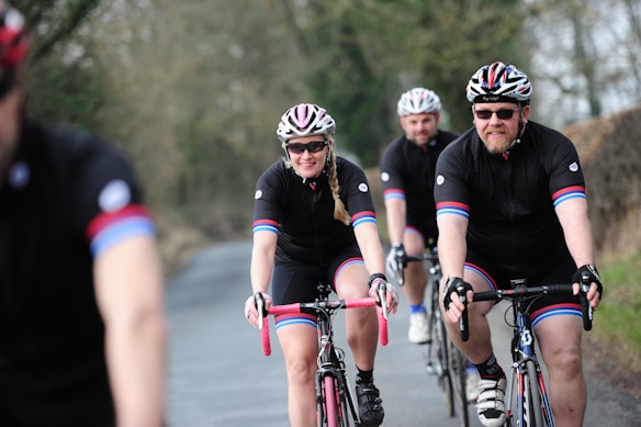 Several cyclists wearing matching black cycling outfits with red and blue stripes are riding on a road through a rural area. They are all wearing helmets and sunglasses. The path they are on is bordered by trees and bushes, creating a natural setting.
