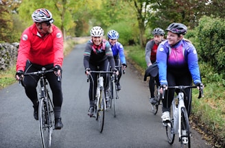 A group of five cyclists are riding on a country road surrounded by lush greenery. They are all wearing helmets and cycling attire, indicating they are on a casual group ride. Their expressions are cheerful, suggesting enjoyment and camaraderie during the activity.